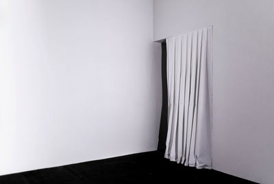 In the corner of a white room there is a dark exit partially covered by a white curtain. 