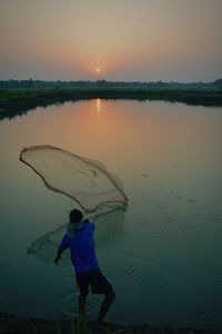 Rear view of fisherman throwing fishnet in lake against sky during sunset