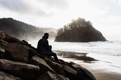 Back view of unrecognizable male tourist sitting on wet stones on beach at seaside in highland area in foggy morning