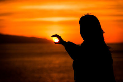 Optical illusion of silhouette woman holding sun at beach during sunset