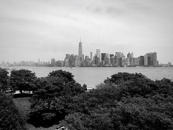 Trees by river and city buildings against cloudy sky