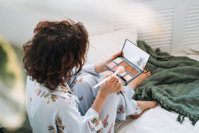 Young woman in blue pajamas applies makeup with face sculpting palette sitting on bed at home