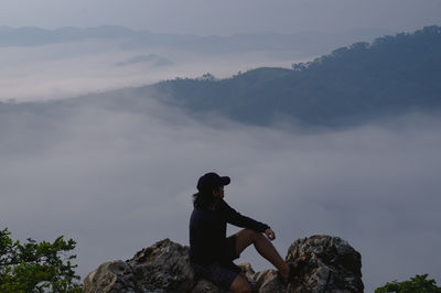 Man sitting on rock looking at mountains against sky