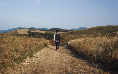 Rear view of woman walking on footpath amidst field against clear sky