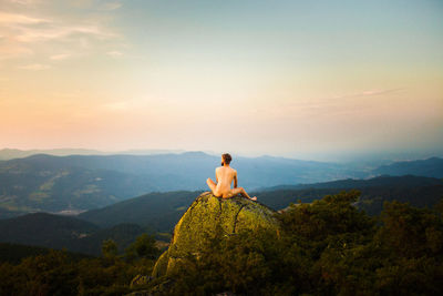 Rear view of naked man sitting on rock against mountains and sky