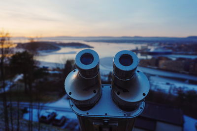 Close-up of coin-operated telescope over town by river during sunset