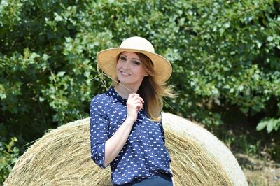 Portrait of beautiful woman holding straw standing by hale bale against trees