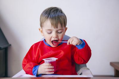 Cute boy eating food while sitting on high chair against wall