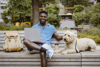 Portrait of smiling man with laptop sitting by labradoodle dog on bench in park