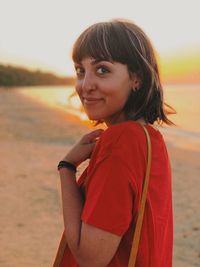 Portrait of smiling young woman standing at beach against sky during sunset