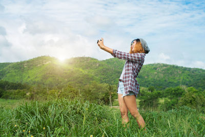 Woman photographing through camera while standing on field against sky