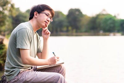 Young man looking away while sitting with book and pen by lake