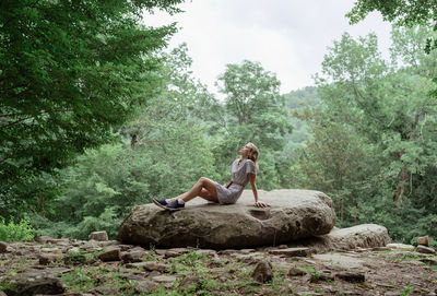 Young woman in summer dress sitting on a big rock in the forest, having rest or meditating