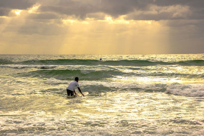 Man with surfboard in sea against sky during sunset