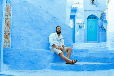 Full length of man sitting on retaining blue wall in town