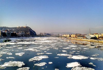 Frozen river by buildings against clear sky during winter