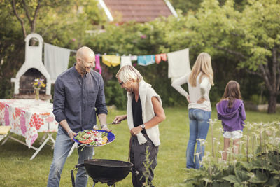 Man and woman cooking vegetables on barbecue grill with girls standing in back yard during garden party