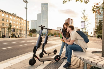 Man showing smart phone to girlfriend while sitting by electric push scooters in city