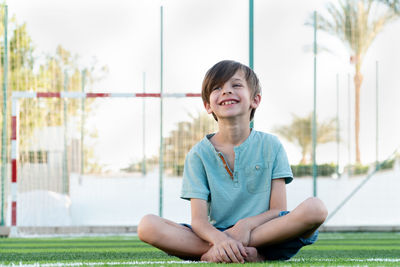 Happy boy sitting on green grass at stadium with crossed legs and looking to distance