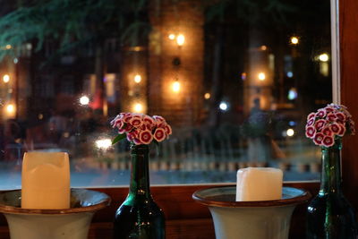 Close-up of candles by flowers in vases against window at night