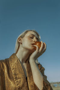 Low angle view of woman with eyes closed eating apple against clear sky