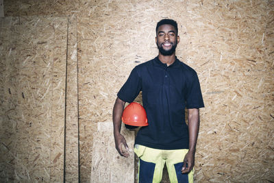 Portrait of confident carpentry student holding hardhat while standing against wooden wall