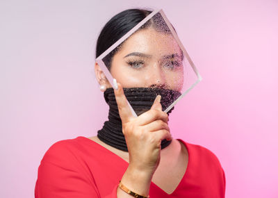 Portrait of young woman with tied mouth holding wet glass against pink background