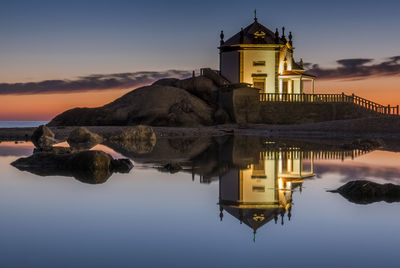 Traditional building by lake against sky during sunset