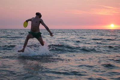 Full length of shirtless man in sea against sky during sunset