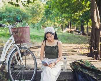 Asian woman in casual wear reading a book on a wooden bridge while resting under the tree