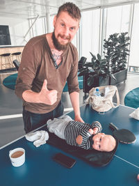 Portrait of father showing thumbs up while standing by son lying on tennis table at home