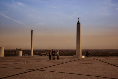 People standing next to sundial on street against sky during sunset