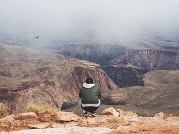 Rear view of man crouching on rock against sky