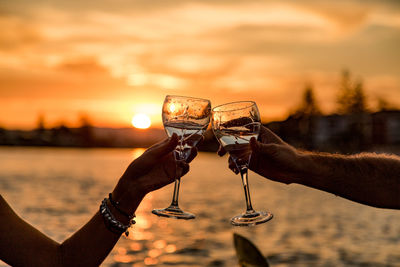 Cropped hands of friends toasting alcoholic drinks by lake against sky during sunset