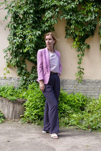 Young blonde stands and straightens hair near the wall of an old building overgrown with wild grapes