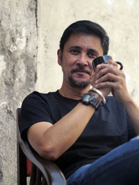 Portrait of man holding mobile phone sitting against wall