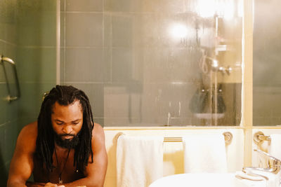 Shirtless man with dreadlocks sitting in bathroom