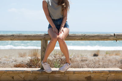 Low section of woman sitting on railing at beach against sky