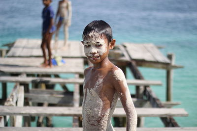 Shirtless boy covered with sand standing on pier over sea