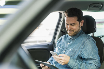 Businessman sitting in car using smartphone and digital tablet