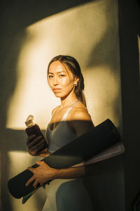 Portrait of woman holding exercise mat while standing against wall