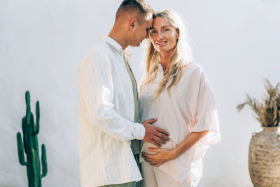 Happy man hugging his pregnant wife outside against a white wall.