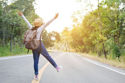 Rear view of playful woman with backpack standing on road amidst trees