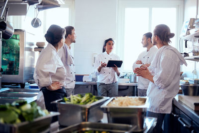 Mid adult female chef holding digital tablet while discussing with team in kitchen