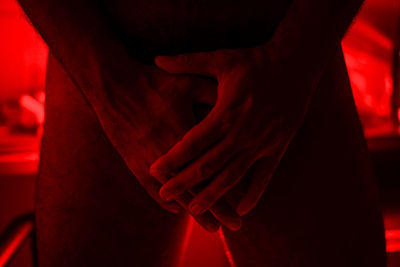 Midsection of naked man gesturing while standing in illuminated red room