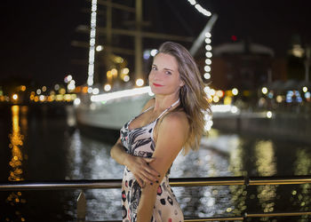 Portrait of beautiful woman standing at riverbank in illuminated city