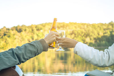 Cropped hands of friends toasting while sitting against lake
