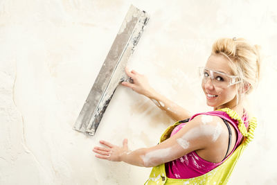 Portrait of young woman in coveralls working with putty knife against white wall