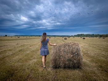 Woman in dress standing on a field near a bale of hay and looking at the dark clouds in the sky