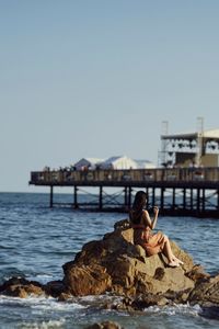 Woman sitting on rock in sea against clear sky